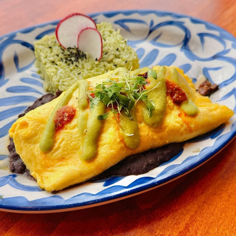 Omelet with black beans, avocado rice, radish slices, and green and red sauces on a blue and white plate.