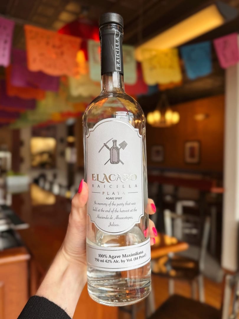 Hand holding a bottle of Elacajo Raicilla agave spirit, with colorful papel picado in the background.